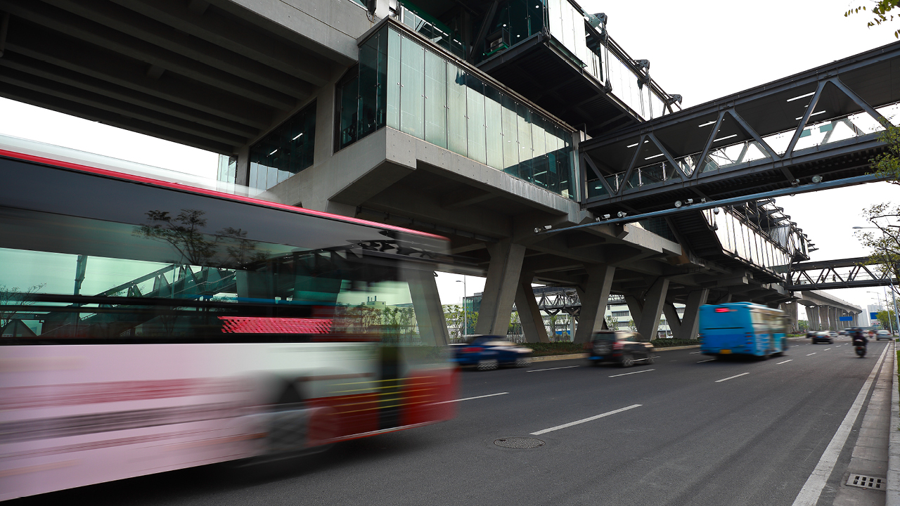 Blurred buses and vehicles move along a city roadway beneath a large elevated transit station and pedestrian walkway.