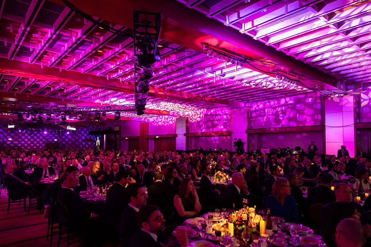 Wide shot of a large ballroom at the Annual Dinner, with more than 1,000 guests seated at round tables under pink and purple stage lighting as a speaker presents on stage beside a screen