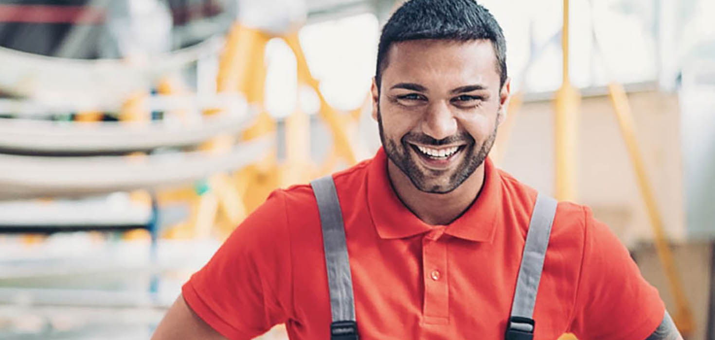 Worker in overalls smiles while standing in front an industrial workplace.