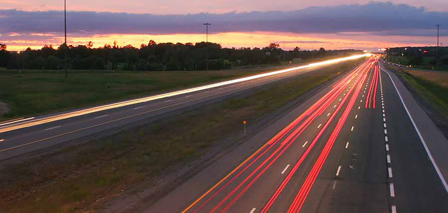 A rural highway at dusk, with streaks of light created by a long exposure indicate where cars have passed.