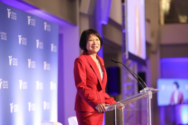 Mayor Olivia Chow stands at a clear podium speaking at an event, wearing a red suit, with a Toronto Region Board of Trade backdrop behind her.