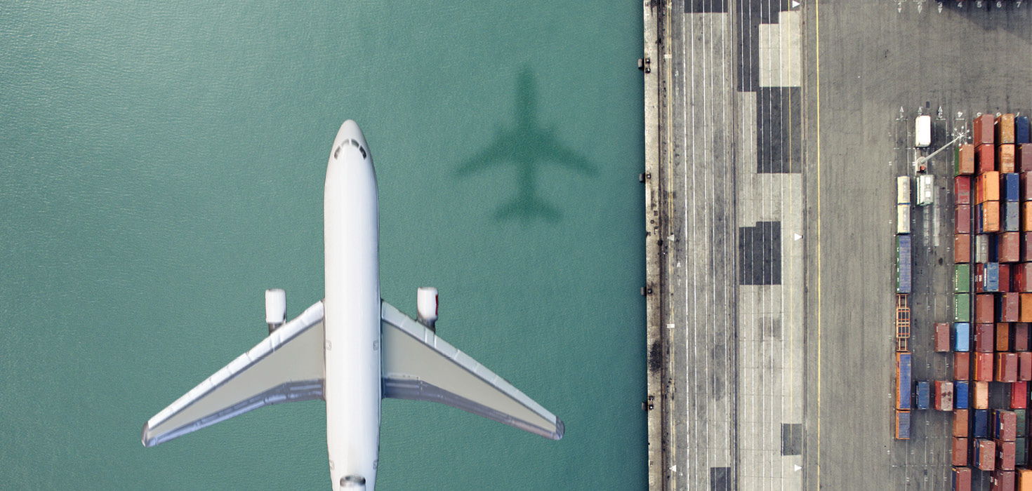 Bird's eye view of an airplane flying over water next to a shipping dock.