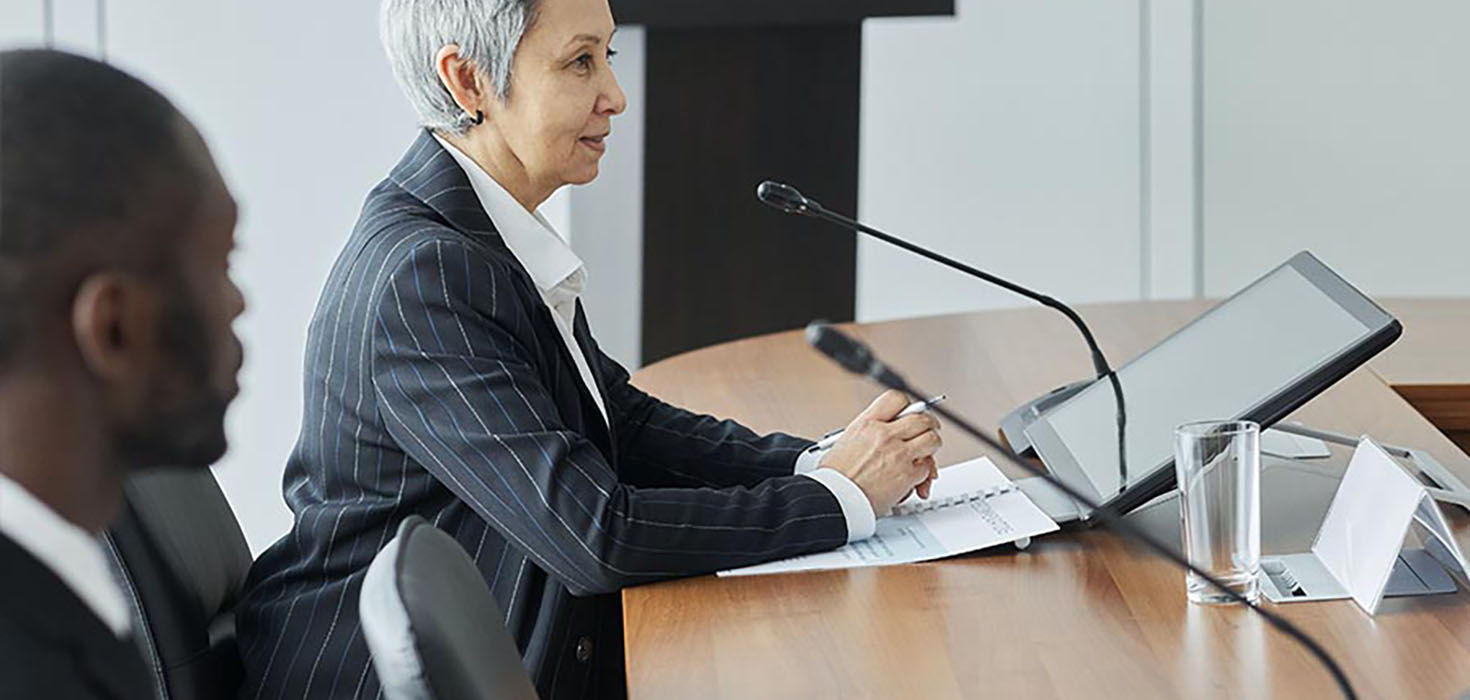 A woman in a suit speaks into a microphone at a board table as a man looks onwards towards her.