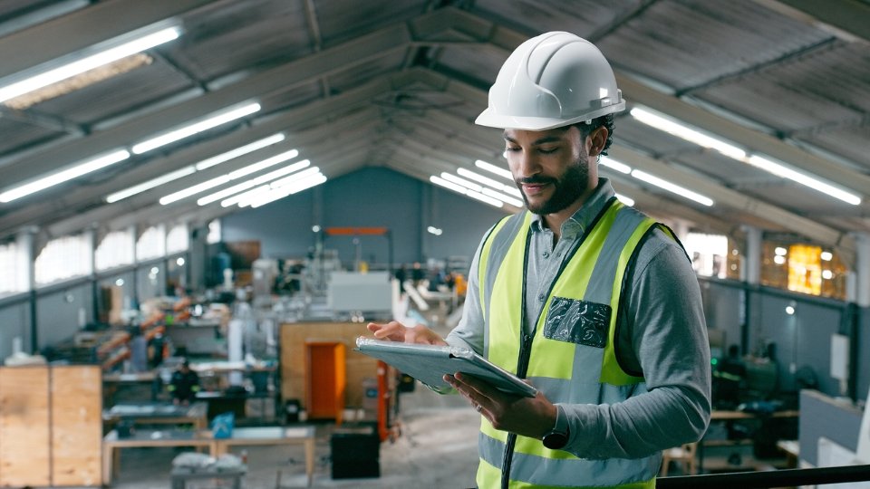 A worker in a safety vest checks a tablet in front of a factory floor