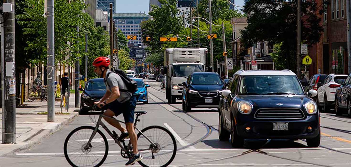 A busy downtown intersection filled with cars and a cyclist. He CN Tower is prominently visible in the background. 